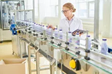Interior of spacious soy milk factory: modern conveyor belt with plastic bottles, concentrated worker wearing white coat controlling production process