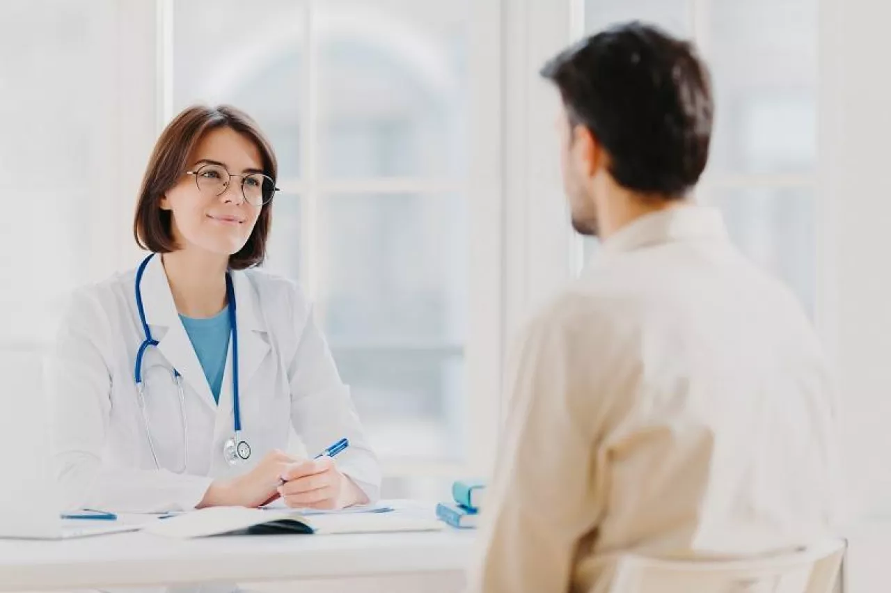 Doctor and patient discuss something, sit at table in clinic. Female cardiologist in eyewear gives medical consultation diagnostic, advice for man how to cure disease, pose in hospital room.