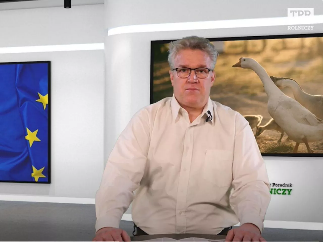 mężczyzna przed mównicą, a za nim dwa ekrany telewizyjne