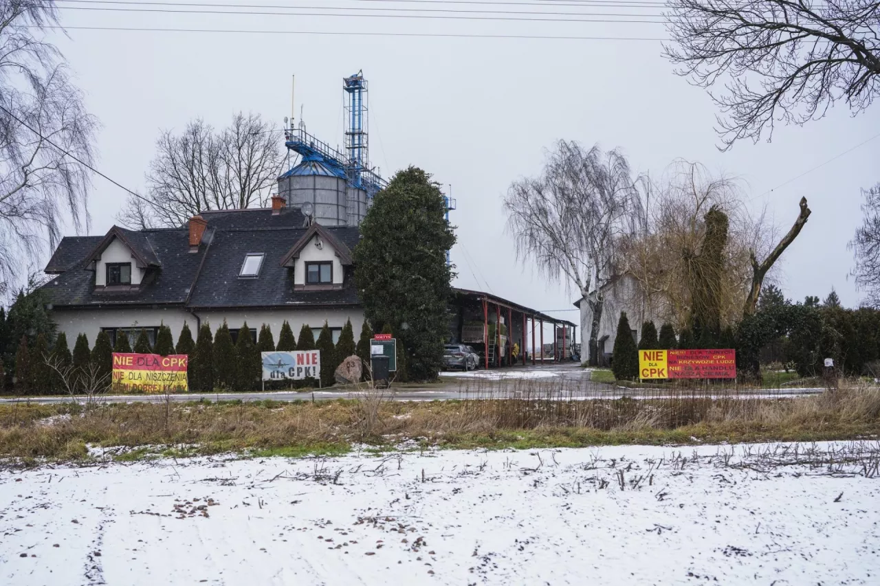 Budynek mieszkalny, a za nim gospodarstwo rolne, a na płocie wywieszone banery sprzeciwiające się budowie CPK