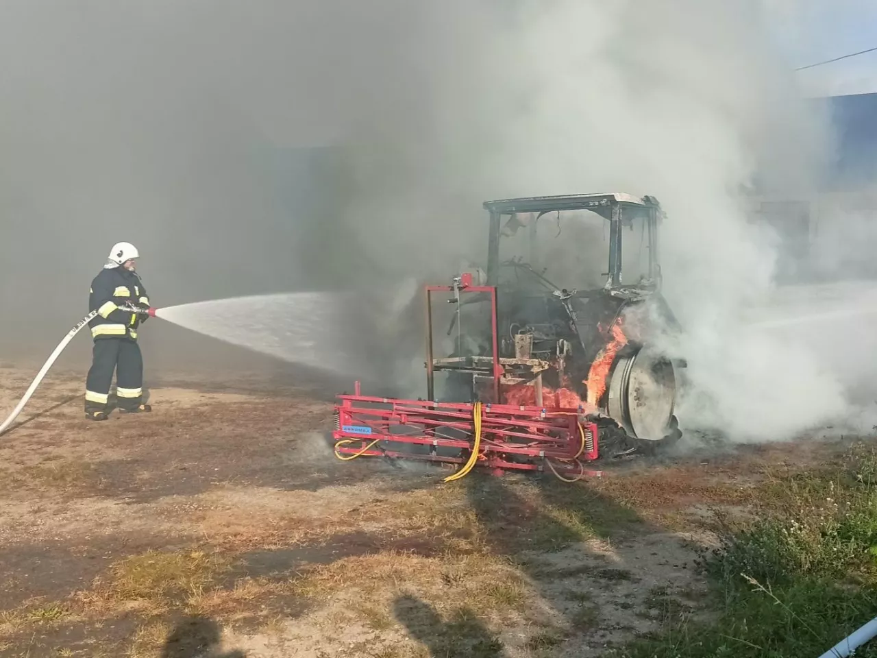 Strażak w czarnym nomexie gasi pożar ciągnika rolniczego z opryskiwaczem