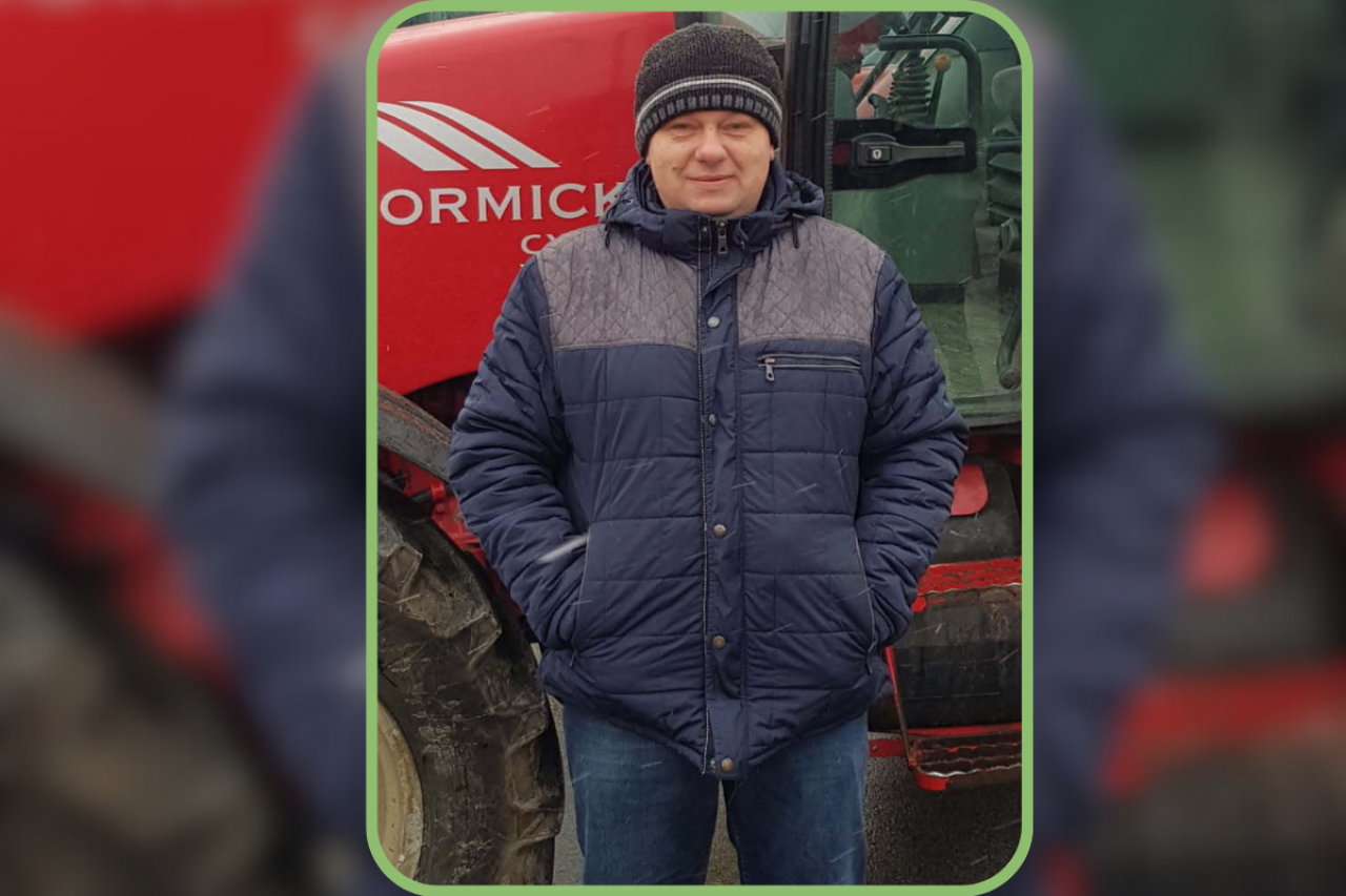 Rolnik w kurtce i czapce stojący przed ciągnikiem rolniczym