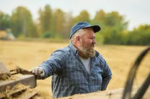 Caucasian middle aged man standing outdoors in field smiling and leaning on farming equipment, wearing cap and gloves, appearing engaged in agricultural work during harvest season