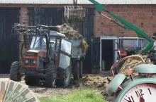 Ciągnik rolniczy przy budynku obory i pryzmie z obornikiem, z podczepionym rozrzutnikiem, a także cyklop, plik banknotów i buzik