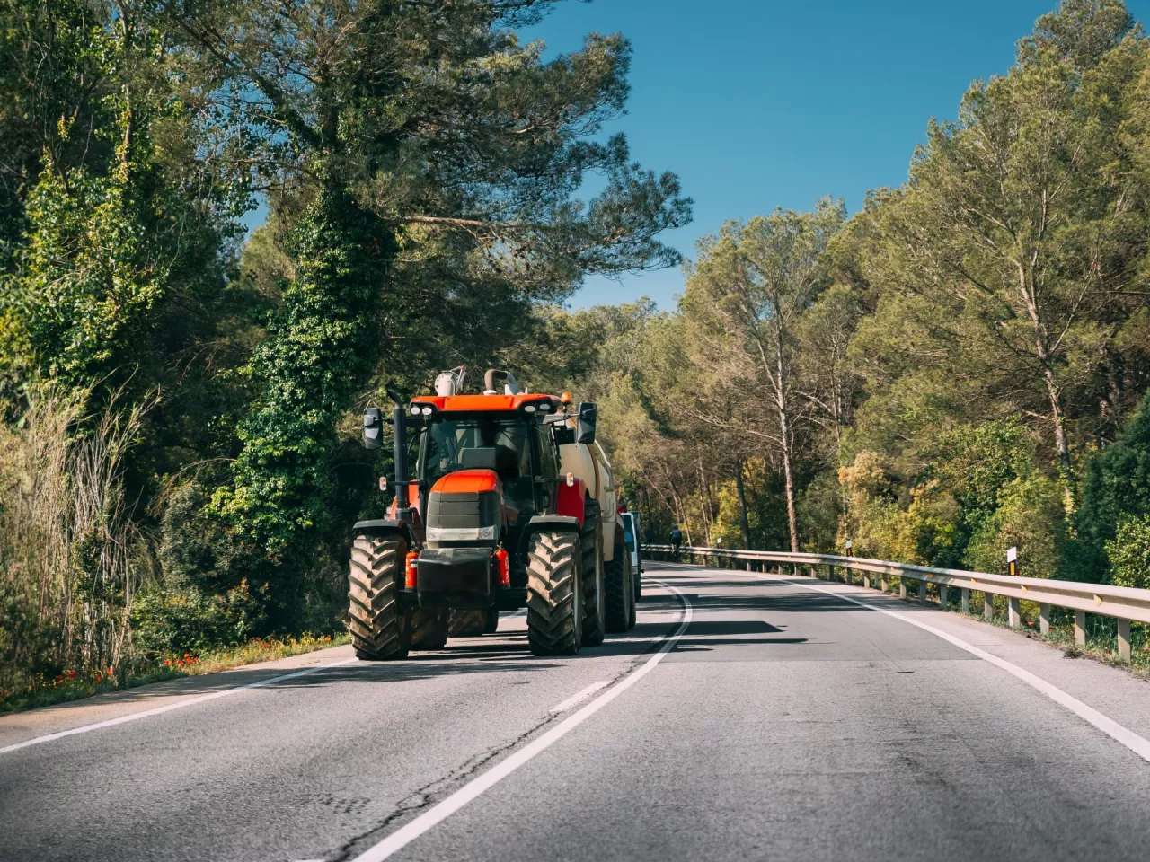 Tractor With Fertilizer Applicator With Tank In Motion On Country Road In Europe. Asphalt Road Against Background Of European Forest Landscape. Agricultural Concept