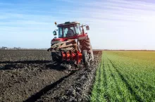 Czerwony ciągnik rolniczy z pługiem obracalnym wykonuje orkę na polu wczesną wiosną. Na pierwszym planie widoczne świeżo zaorane skiby, obok rzędy zielonych roślin ozimych. Scena przedstawia typową pracę rolnika w okresie przygotowań do nowego sezonu.