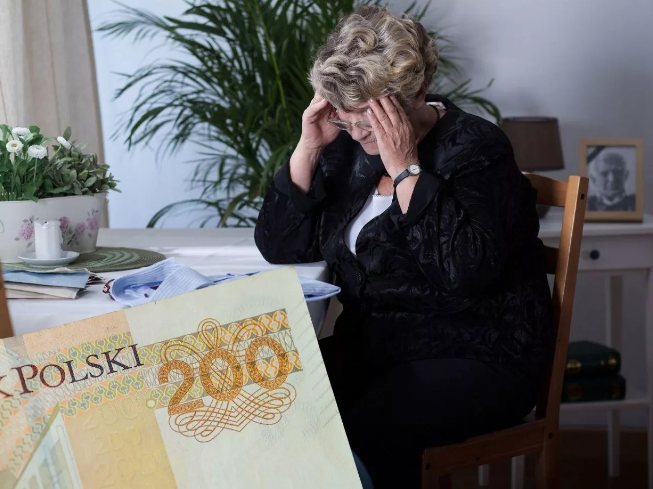 starsza kobieta, wdowa siedzi przy stole, na stole męska koszula, w lewym dolnym rogu kawałek banknotu 200 zł